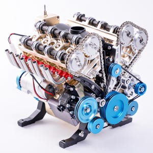 Könnte beinhalten: Ein detailliertes V-Motormodell mit goldfarbenem Motorblock, silbernen Steuerketten und metallisch blauen Riemenscheiben. Die Baugruppe umfasst rote Ansaugstutzen, schwarze Strukturkomponenten und eine Reihe silberner Auspuffrohre.