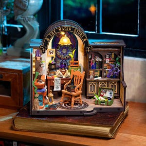 May include: A miniature wooden diorama titled The Magic Study featuring a desk, a chair with a black hat, a bookshelf filled with books, a glowing lamp, a cauldron, a crystal ball, a broom, and various small accessories in a detailed room setting.