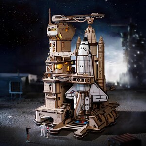 May include: A detailed wooden model of a space station and a space shuttle. The model features multiple levels, a launch pad, and a crane. The shuttle is white with black accents and the word "SPACE" on the side. The model is set against a starry night backdrop.