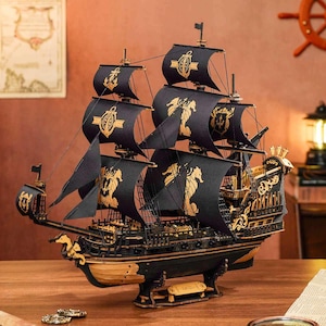 May include: A black wooden model ship with gold accents and black sails. The sails have gold seahorse and crown designs. The ship has a detailed hull and intricate carvings.