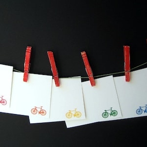May include: Seven blank white cards with a bicycle design printed on each card are hanging on a black background with red clothespins. The bicycles are in different colors: red, blue, green, yellow, and orange.