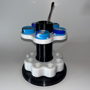 Puede incluir: Un organizador impreso en 3D en blanco y negro con una columna central negra y una base blanca. El organizador tiene seis ranuras para contenedores y un soporte para una herramienta. Los contenedores son azules, blancos y negros.