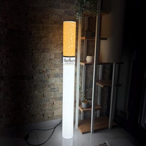 May include: A novelty floor lamp in the shape of a cigarette. The lamp has a white cylindrical body with a yellow-orange top. The text "Marlboro Lights" is printed on the side. The lamp is plugged into a wall socket.