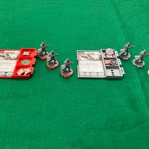 May include: A tabletop game set featuring six gray plastic miniature figures of soldiers in long coats, two rectangular plastic trays in red and gray, and several circular game tokens with various symbols printed on the cards and tokens.
