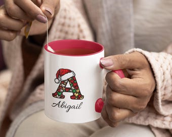 Op de afbeelding: Witte keramische mok met een rode binnenkant en handvat. De mok heeft een kerstthema-ontwerp met de letter "A" versierd met feestelijke patronen en een kerstmuts. De naam "Abigail" staat eronder.