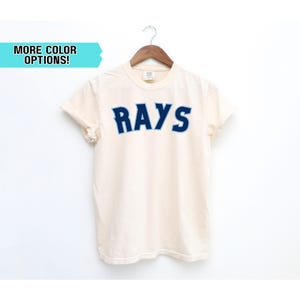 May include: Cream-colored t-shirt with the word "RAYS" in blue block letters. The shirt is hanging on a wooden hanger against a white background. Text on the left says "MORE COLOR OPTIONS!"