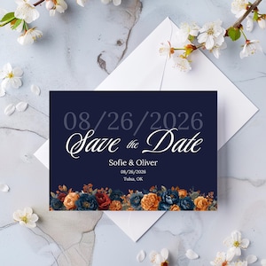 Może przedstawiać: Granatowa karta "Save the Date" z datą 08/26/2026 w szarym i białym skrypcie. Karta zawiera imiona Sofie & Oliver oraz lokalizację Tulsa, OK. Kwiatowe akcenty w kolorze pomarańczowym, niebieskim i czerwonym zdobią dół. Karta spoczywa na białej kopercie, z białymi kwiatami rozsianymi wokół.