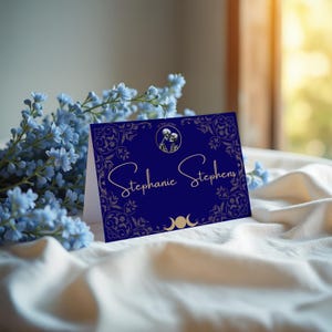 May include: A navy blue card with gold filigree and the name "Stephanie Stephens" in elegant script. The card features a small circular image of two skeletons and crescent moons. Blue flowers are in the background.