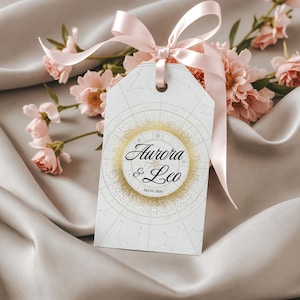 Könnte beinhalten: Ein weißes Geschenk-Etikett mit einem goldenen Himmelsdesign und den Namen "Aurora & Leo" in eleganter Schrift. Das Etikett ist mit einem rosa Band gebunden und liegt auf einer Satin-Oberfläche, mit rosa Blumen im Hintergrund.