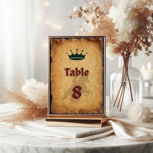 May include: A gold-framed table number card with a parchment-style background. The card features a green crown, the word "Table" in burgundy, and the number "8" with a knight on horseback. The card is displayed on a gold stand.