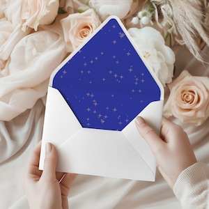 May include: A white envelope is held open, revealing a deep blue interior adorned with a pattern of small, light-colored stars. The envelope is set against a backdrop of soft, cream-colored fabric and delicate flowers.