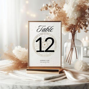 May include: A gold-framed table number card with the word "Table" in elegant script above the number "12". The number is set against a zodiac wheel design. The names "Aurora & Leo" and the date "24th September 2026" are printed below.
