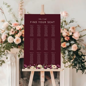 May include: A burgundy seating chart with the text "Find Your Seat" and names listed under table numbers. The chart is displayed on a wooden easel and decorated with floral arrangements.
