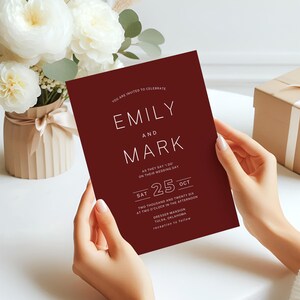 May include: A burgundy wedding invitation with white text, held by hands. The invitation reads "Emily and Mark" with the date "25 Oct". A bouquet of white flowers and a gift box are in the background. The invitation is for a wedding.