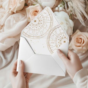 May include: A white envelope with a gold zodiac design on the inside. The envelope is held open, revealing the intricate celestial map. The background features soft pink and white flowers, creating a delicate and elegant aesthetic.