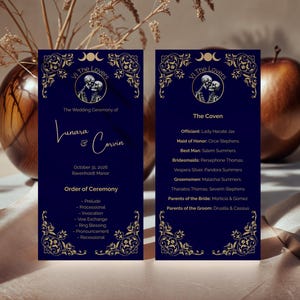 May include: Two dark blue wedding ceremony cards with gold accents. The cards feature the text "Vi The Lovers" and "Lunara & Corvin". The cards list the order of ceremony and the wedding party details. The cards are set against a neutral background.