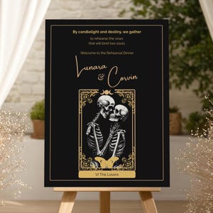 May include: A black rehearsal dinner sign on an easel. The sign features gold text and a gold-framed illustration of two skeletons embracing. The text reads "By candlelight and destiny, we gather...Welcome to the Rehearsal Dinner Lunara & Corvin". The bottom reads "VI The Lovers".