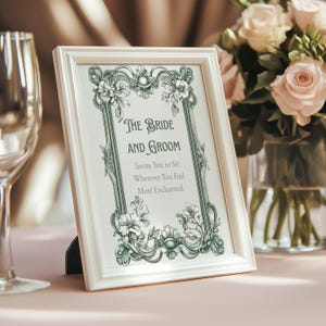 May include: A white framed sign with the text "The Bride and Groom" and floral embellishments. The sign invites guests to sit wherever they feel most enchanted. A vase of pink roses is in the background.