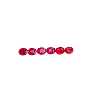 May include: Six oval-cut, vibrant red gemstones are arranged horizontally against a white background. The gems have a polished, reflective surface, showcasing their rich color and clarity. These gemstones could be rubies, garnets, or similar precious stones.