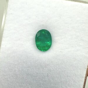 May include: Oval-cut emerald gemstone, a vibrant green hue, set against a white background. The gemstone is displayed in a clear plastic case, showcasing its polished facets and natural inclusions. A close-up view highlights the gem's clarity and color.