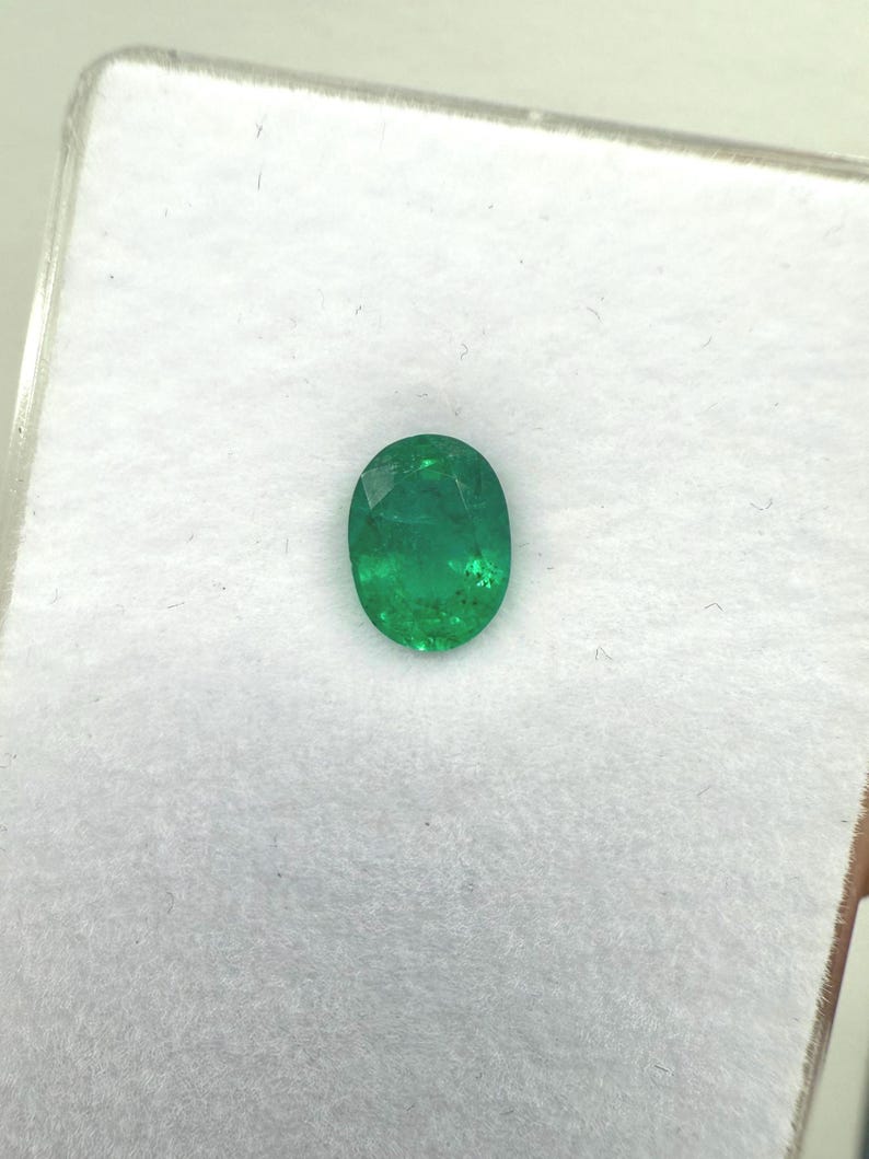 May include: Oval-cut emerald gemstone, a vibrant green hue, set against a white background. The gemstone is displayed in a clear plastic case, showcasing its polished facets and natural inclusions. A close-up view highlights the gem's clarity and color.