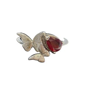 May include: A silver ring featuring a pear-shaped, deep red gemstone. Two detailed butterfly accents flank the gemstone, adding a delicate touch. The ring's band is a simple, polished silver, creating a contrast with the gemstone and butterfly design.