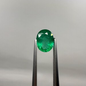 May include: A faceted, oval-cut emerald gemstone held by a pair of tweezers. The emerald is a vibrant green color and appears to be well-polished, reflecting light. The tweezers are silver-colored and have a textured grip.