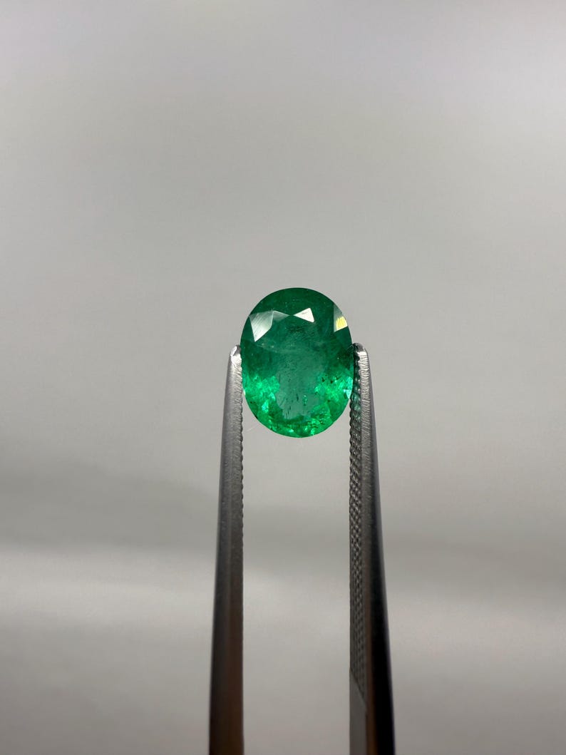 May include: A faceted, oval-cut emerald gemstone held by a pair of tweezers. The emerald is a vibrant green color and appears to be well-polished, reflecting light. The tweezers are silver-colored and have a textured grip.