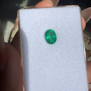 May include: A vibrant green oval-cut emerald gemstone displayed in a white velvet-lined box. The emerald has a rich, saturated color and is set against a neutral background, highlighting its clarity and brilliance. The box is held in a person's hand.