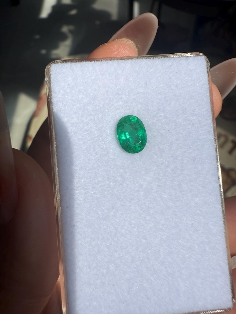 May include: A vibrant green oval-cut emerald gemstone displayed in a white velvet-lined box. The emerald has a rich, saturated color and is set against a neutral background, highlighting its clarity and brilliance. The box is held in a person's hand.