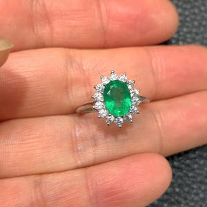 May include: A silver ring featuring an oval-cut emerald gemstone, surrounded by a halo of small, round, white gemstones. The emerald is a vibrant green color, and the ring is set against a neutral background.