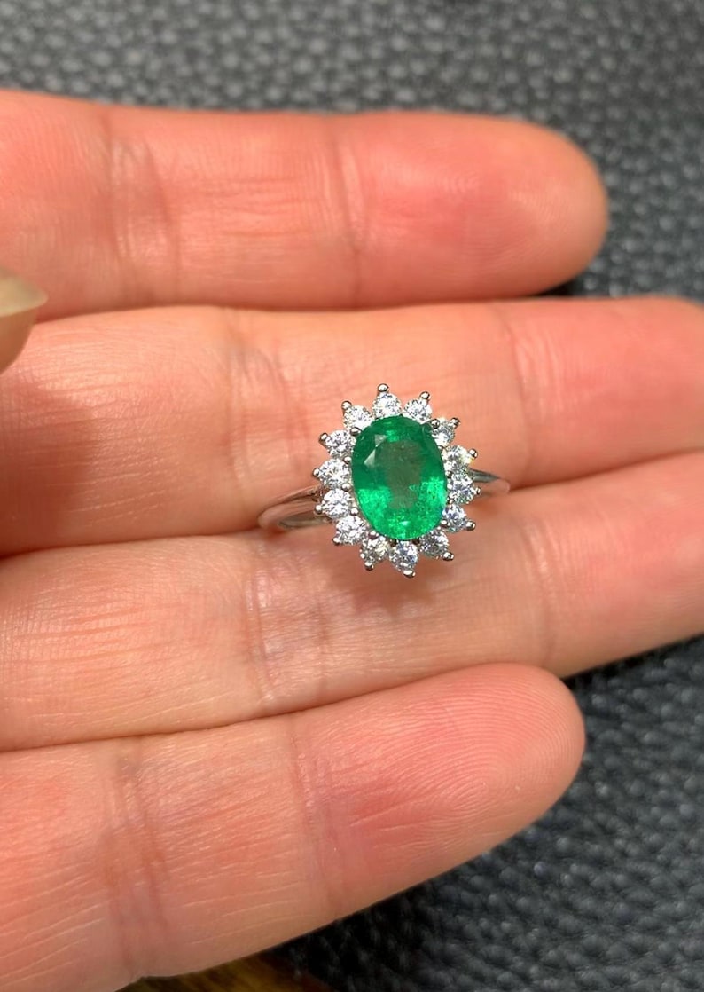 May include: A silver ring featuring an oval-cut emerald gemstone, surrounded by a halo of small, round, white gemstones. The emerald is a vibrant green color, and the ring is set against a neutral background.