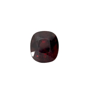May include: A faceted, cushion-cut gemstone with a deep red color. The gem has a square shape with rounded corners and a polished surface, reflecting light. The gemstone is set against a white background.