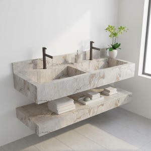May include: A modern double sink made of light gray stone with brown veining. The sink has two basins, each with a dark faucet. A lower shelf holds folded white towels and a tray with soap and a stone. A small plant sits on the right.