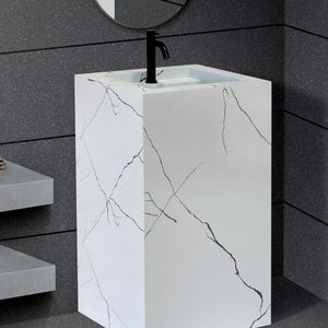 May include: A white pedestal sink with black veining, a black faucet, and a built-in basin. The sink is rectangular and stands on a matching base. The background features a dark gray wall and a round mirror.