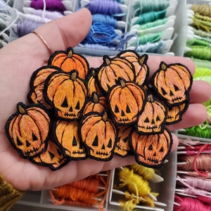 May include: A collection of embroidered pumpkin patches with black stitched details, including eyes, nose, and mouth. The patches are orange and yellow with a slight gradient effect.