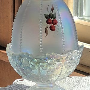 May include: An iridescent glass egg-shaped candle holder with a floral base and a beaded pattern. The top features a hand-painted design of red berries and green leaves. The candle holder is sitting on a white lace doily.