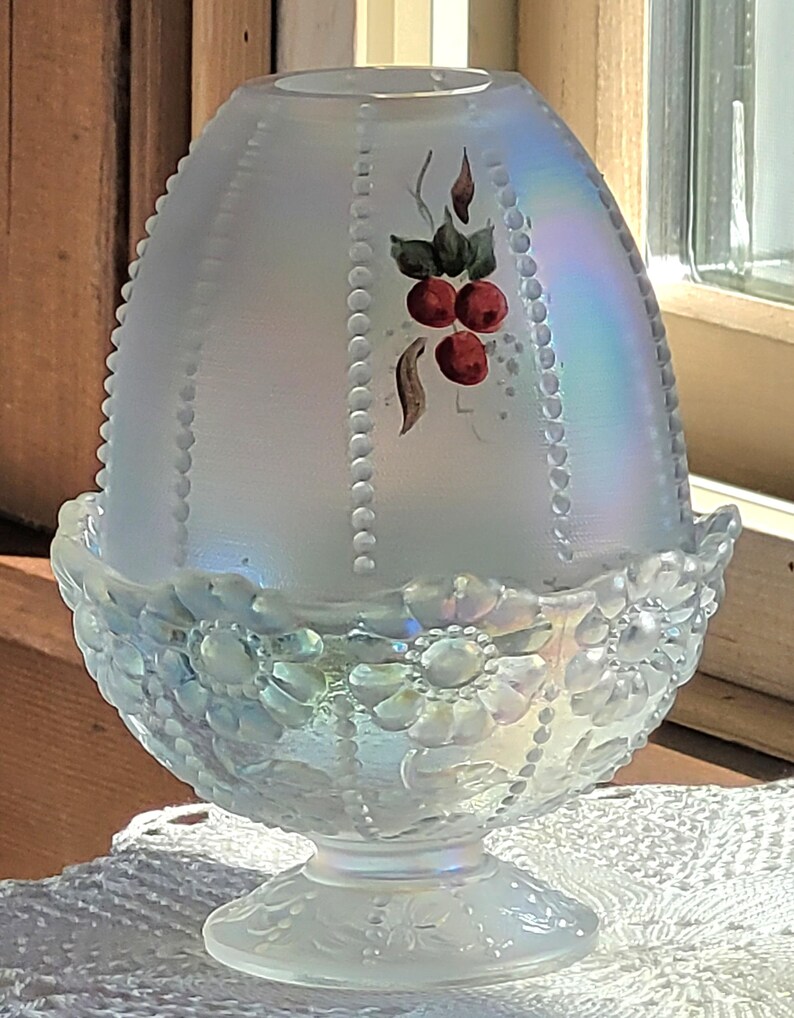May include: An iridescent glass egg-shaped candle holder with a floral base and a beaded pattern. The top features a hand-painted design of red berries and green leaves. The candle holder is sitting on a white lace doily.