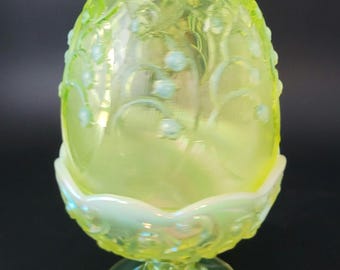 May include: A bright yellow, translucent glass egg-shaped vessel with an iridescent finish. The surface features embossed floral patterns and sits atop a scalloped, pedestal-style base with a matching yellow and iridescent glass finish.