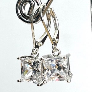 May include: A pair of silver leverback earrings with square, clear cubic zirconia stones. The earrings are displayed against a white background, showcasing their sparkling facets and elegant design. The leverback closure is visible.