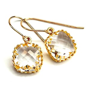 May include: A pair of gold-colored drop earrings with clear, square-cut gemstones. The gemstones are set in a gold-colored, textured bezel. The earrings have a simple hook closure.
