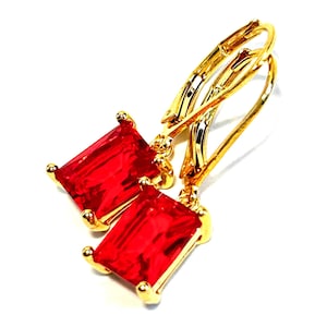 May include: A pair of gold-colored leverback earrings with rectangular, vibrant red gemstones. The earrings are set against a white background, highlighting the contrast between the gold metal and the red stones.