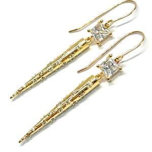 May include: A pair of gold-colored earrings with a unique design. Each earring features a long, textured spike adorned with small, clear gemstones. A square, clear gemstone sits above the spike, connected to a curved ear wire.