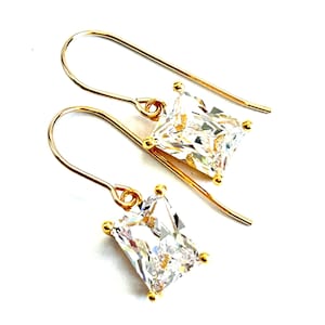 May include: A pair of gold-colored drop earrings with rectangular, clear, faceted stones. The earrings have a simple hook design and small gold accents at each corner of the stones. The background is plain white.