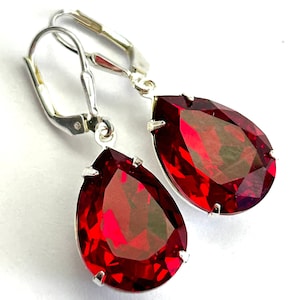 May include: A pair of teardrop-shaped earrings with faceted, deep red gemstones. The earrings have a silver-colored metal setting and lever-back closures. The gemstones are a vibrant, saturated red color, catching the light.