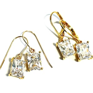 May include: Two pairs of gold-toned earrings featuring rectangular, clear, faceted stones set in four-prong settings. One pair has simple hook wires, while the other pair features lever-back closures, both showcasing brilliant, reflective, clear crystals.