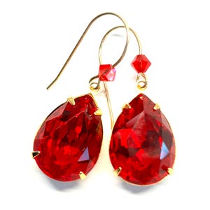 May include: A pair of teardrop-shaped red crystal earrings with gold-colored metal accents. The earrings have a faceted design, catching the light. Small red crystal beads are attached to the gold hooks.