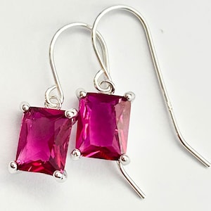 May include: A pair of silver drop earrings with rectangular, magenta-colored gemstones. The gemstones are set in a silver frame with small, round accents at each corner. The earrings have a simple, elegant design.