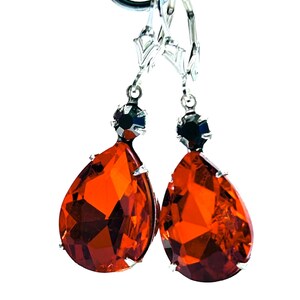 May include: A pair of silver-toned drop earrings with large, faceted, teardrop-shaped orange-red gemstones. Each earring has a small, dark, round stone at the top and a lever-back closure. The earrings are displayed against a white background.