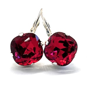May include: A pair of leverback earrings featuring large, faceted, square-cut, ruby-red crystals. The crystals are set in a silver-tone metal. The earrings are displayed against a white background.